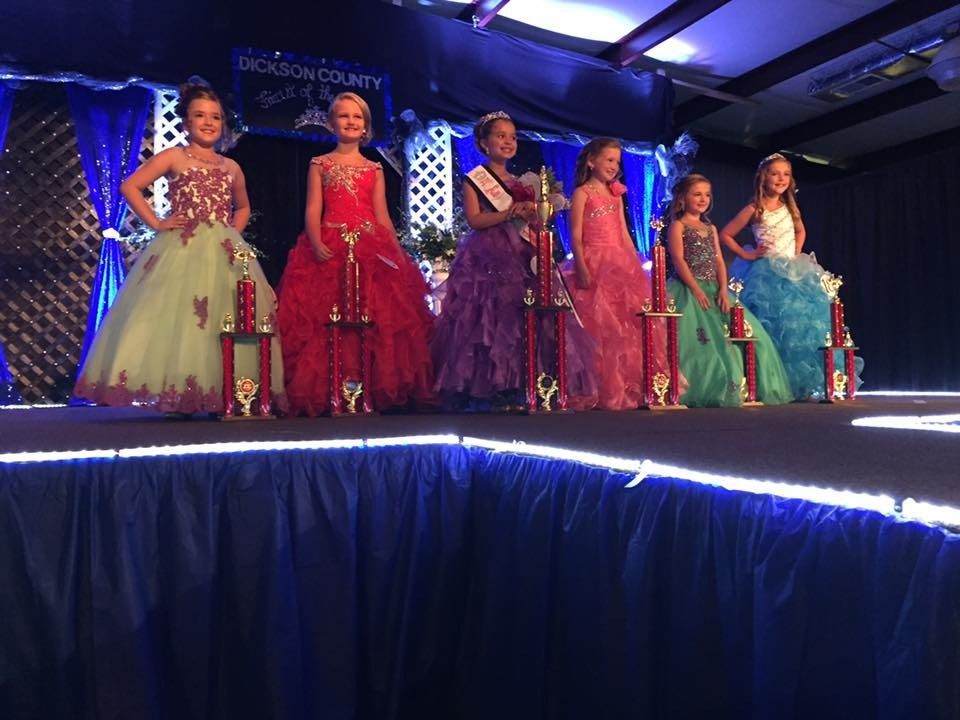 Six children in colorful formal dresses stand on a lit runway stage during a fashion show.