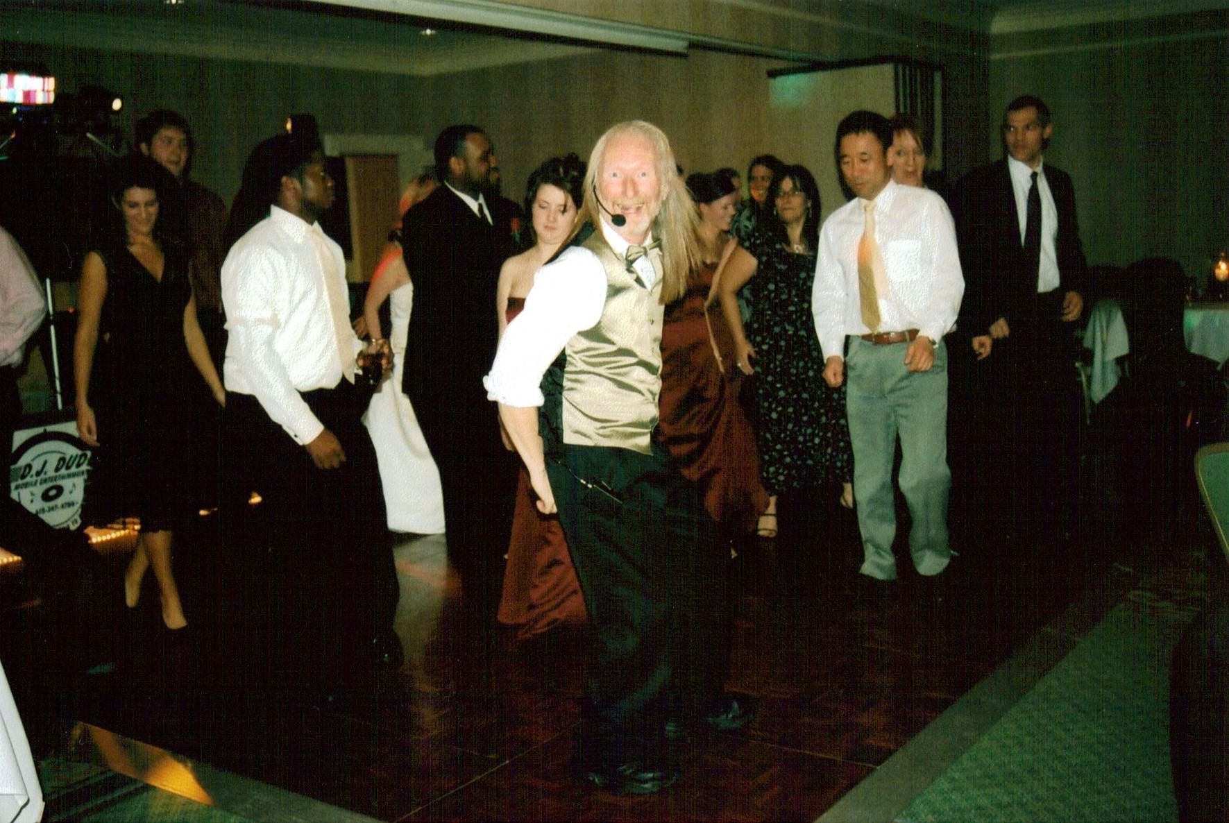 People dancing at a dimly lit party, with one person in a white wig and vest in the center.