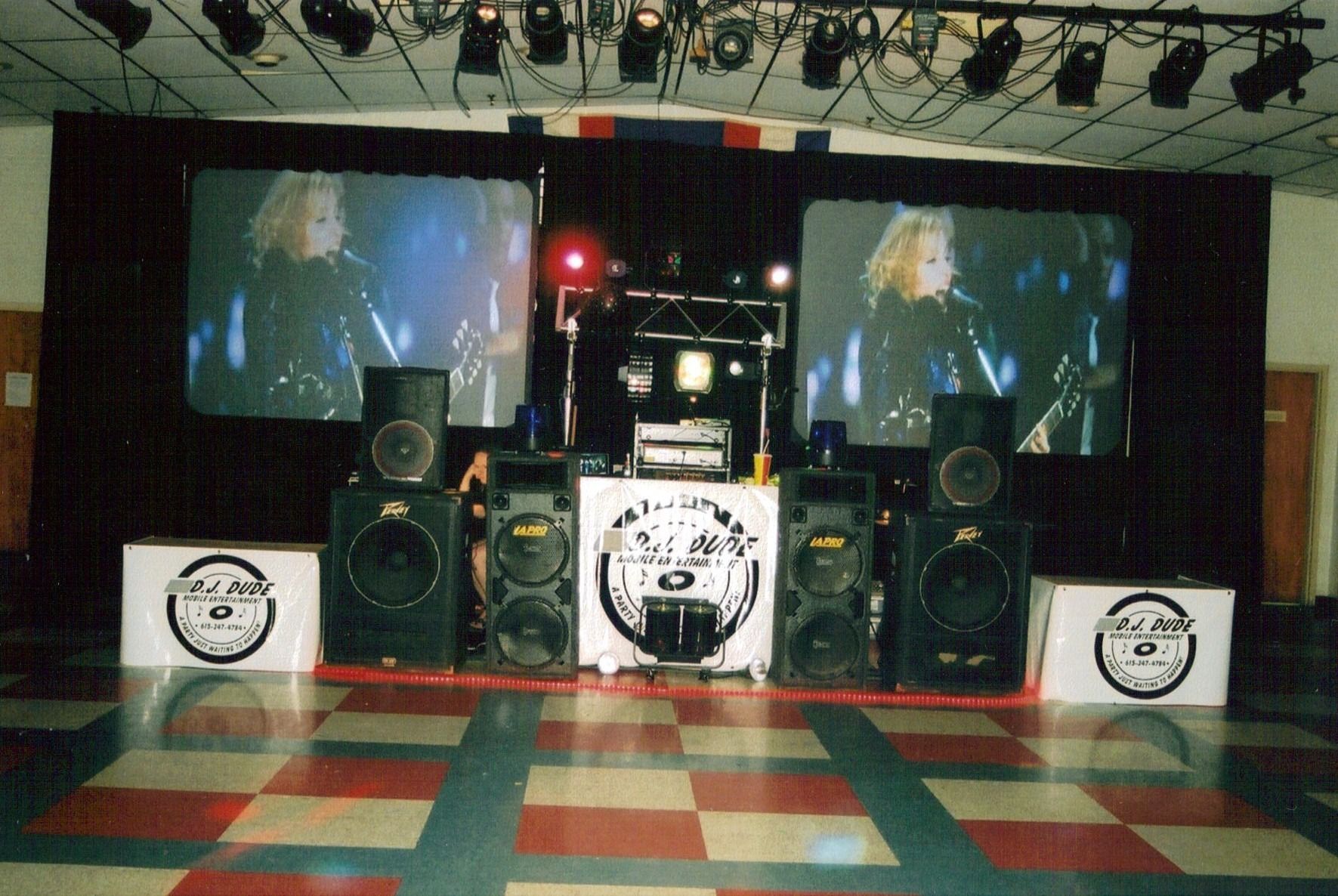 Stage with large speakers, two screens, and a checkered dance floor in a club-like venue