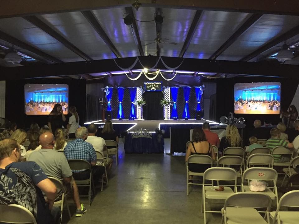Audience seated in a hall facing a blue-lit stage with screens and a podium