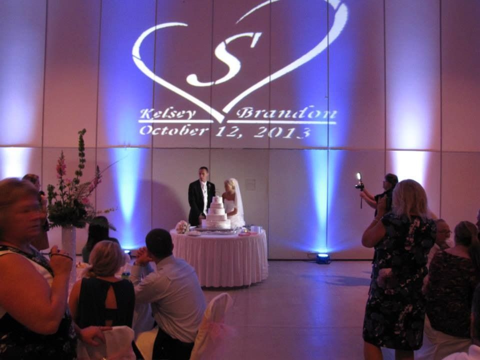 Wedding couple at reception table under monogrammed wall projection, with guests taking photos nearby.