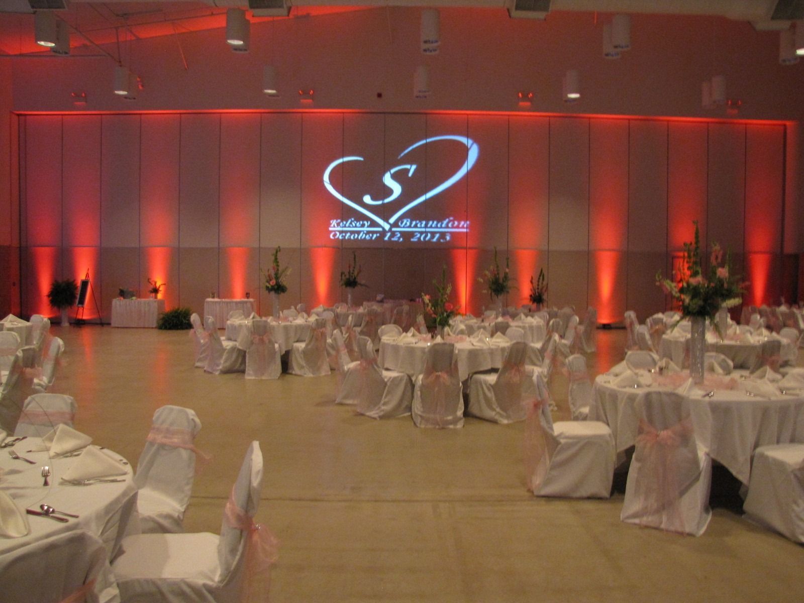 Red-lit banquet hall set for a formal event with round tables, white chairs, and a heart logo on the stage backdrop