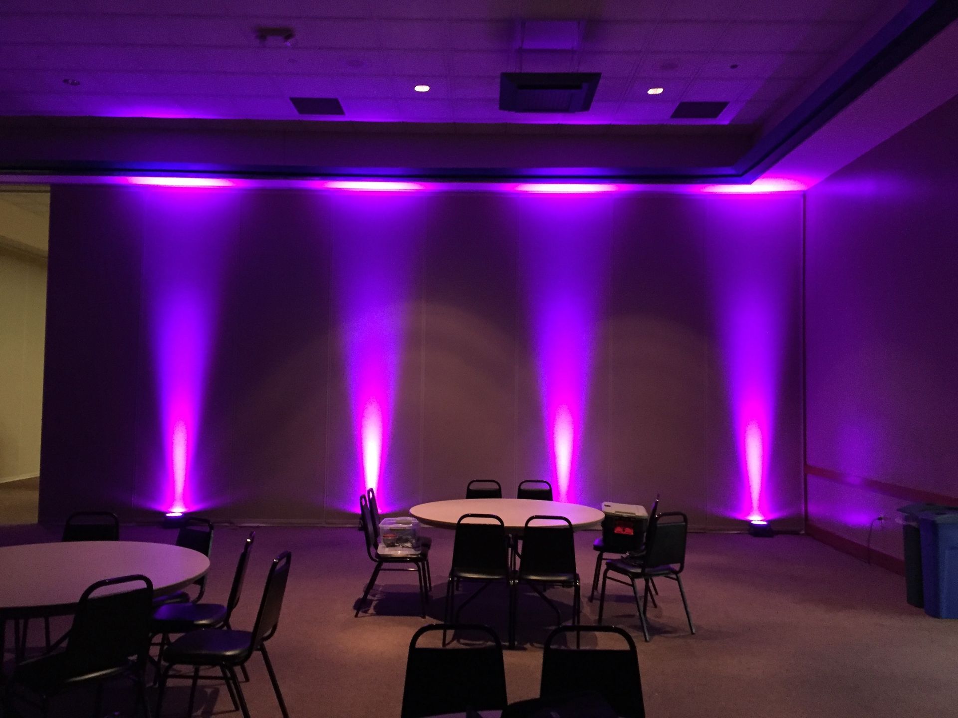 Purple uplighting illuminates an empty banquet room with round tables and chairs.