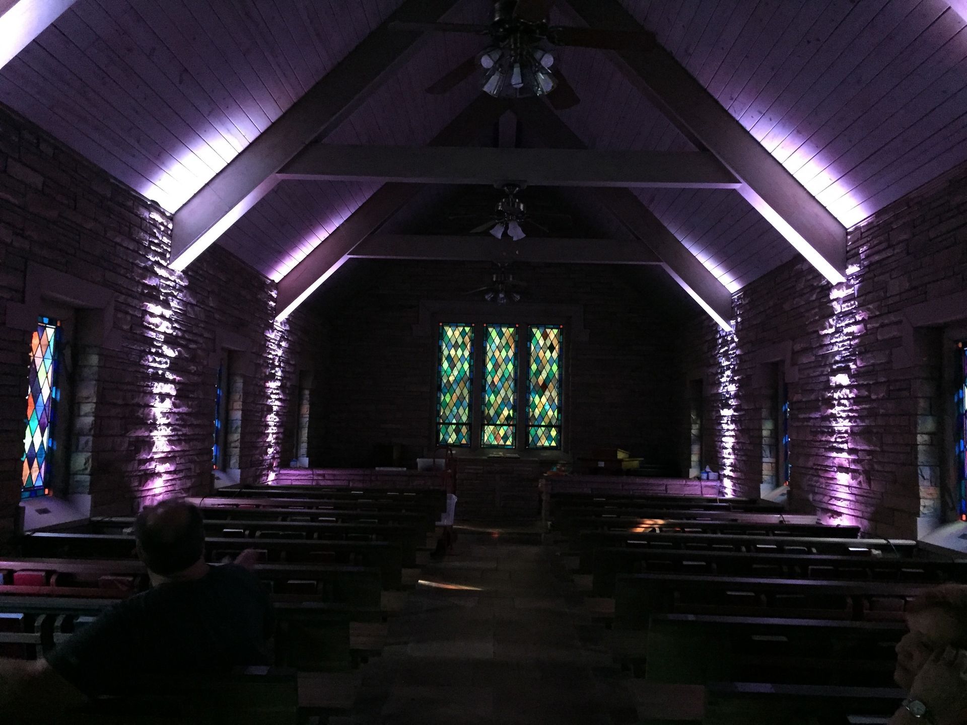Dim church interior with purple uplighting, wooden pews, and stained-glass windows at the front.
