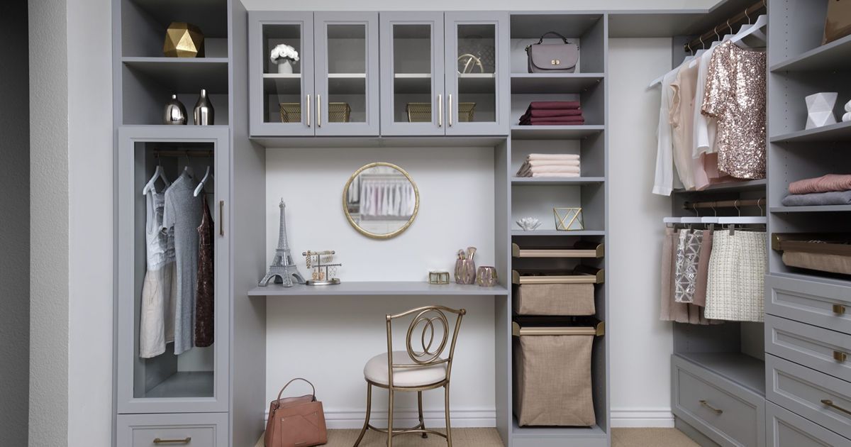 Gray walk-in closet with built-in desk, glass-front cabinets, and clothing racks; neutral tones.