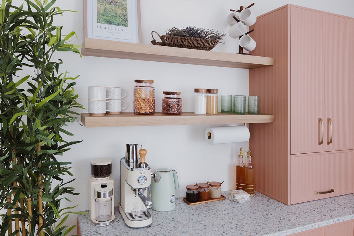 Pink kitchen with open shelves holding jars, cups, and cabinets. A coffee maker sits on the counter near a tall plant.