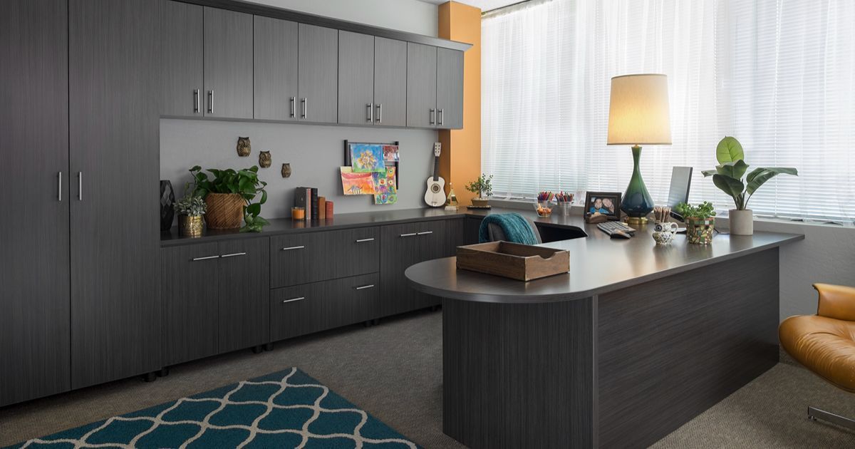 Office with gray cabinets, desk, blue rug, and plants. Window with blinds and a lamp.