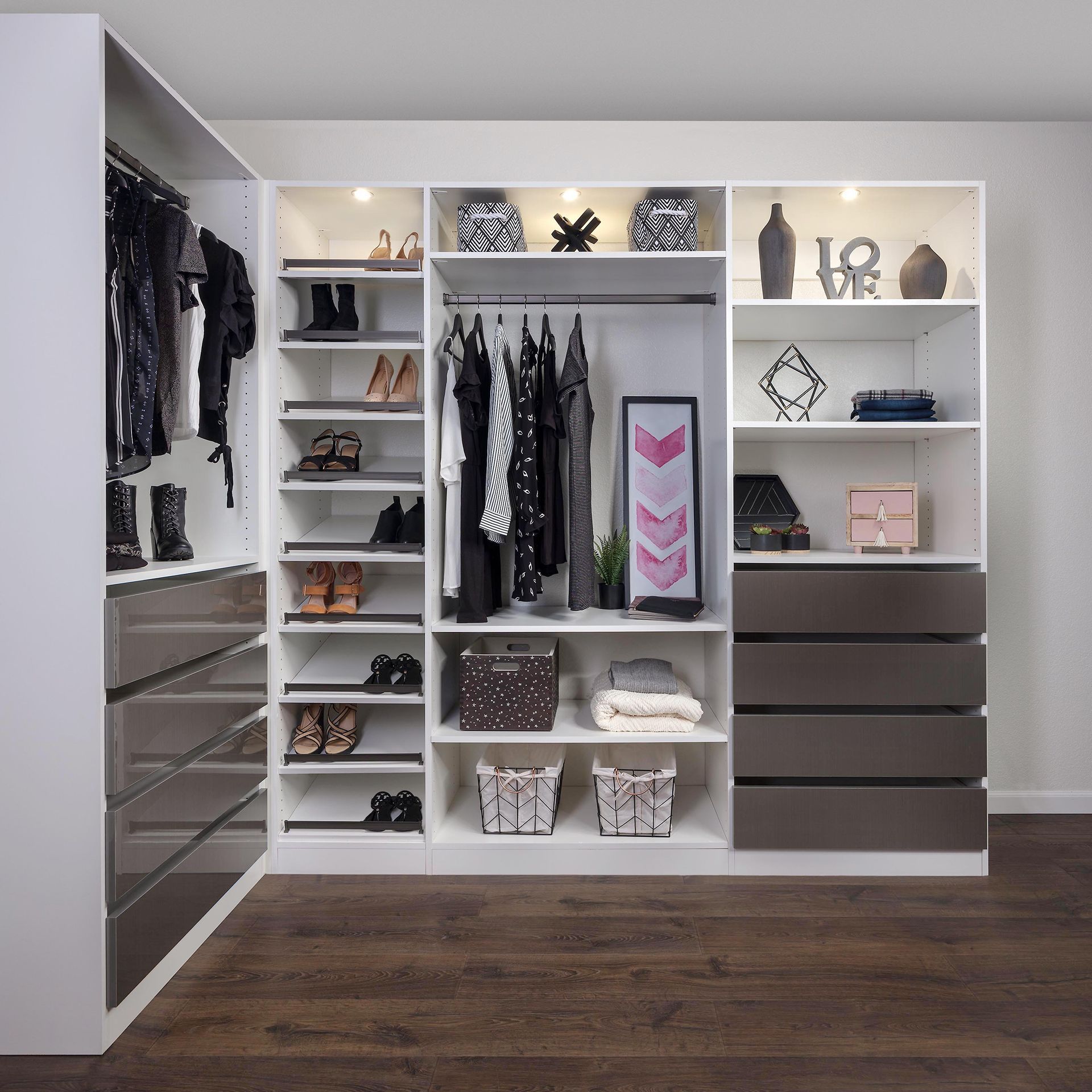 A modern, organized walk-in closet with white shelves, drawers, and hanging space, with dark brown accents and clothes.