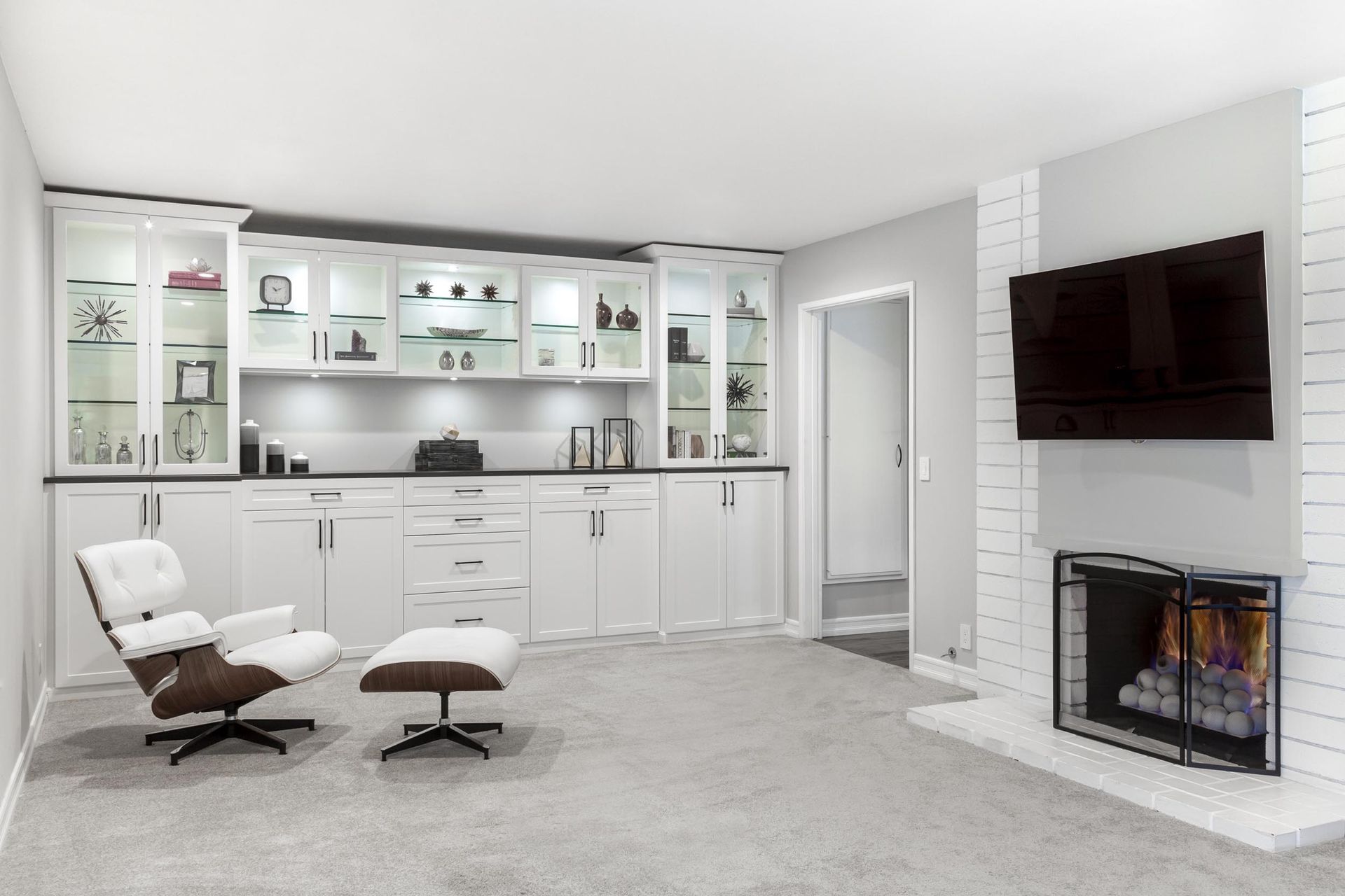 White built-in cabinets with a TV over a brick fireplace, an armchair, and an ottoman in a living room.