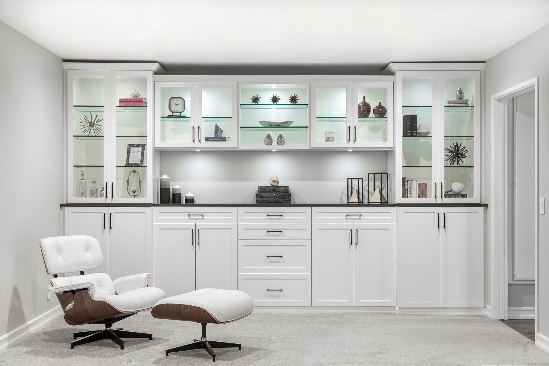 White built-in cabinetry with glass-door uppers, beneath a gray countertop. A chair and ottoman sit in front.