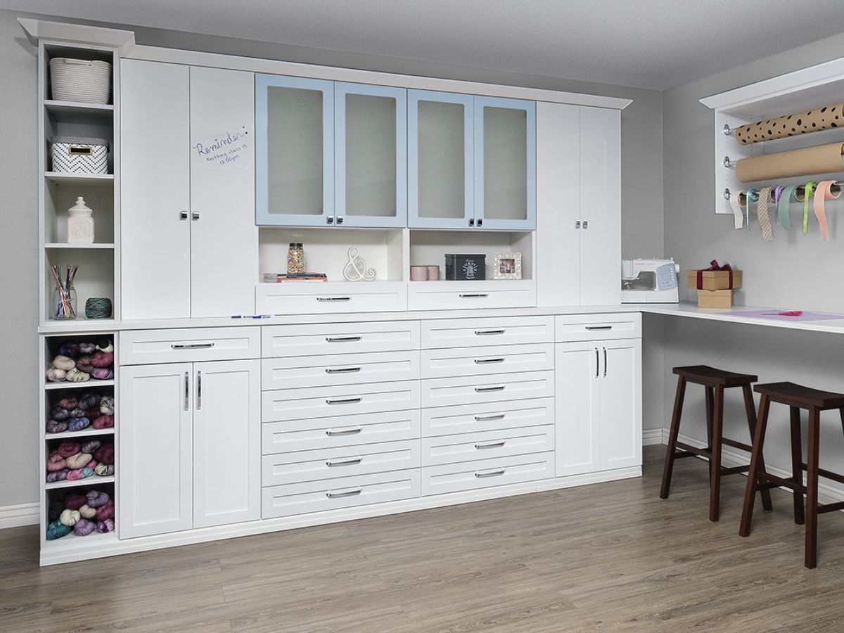 White craft room storage cabinets with drawers, shelves, and blue-tinted glass-door upper cabinets.