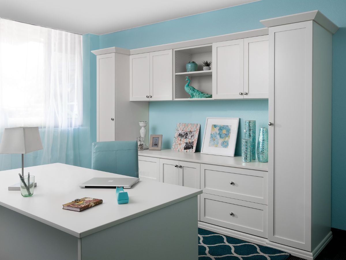 White desk and cabinets in a room with blue walls, a teal chair, and patterned rug.