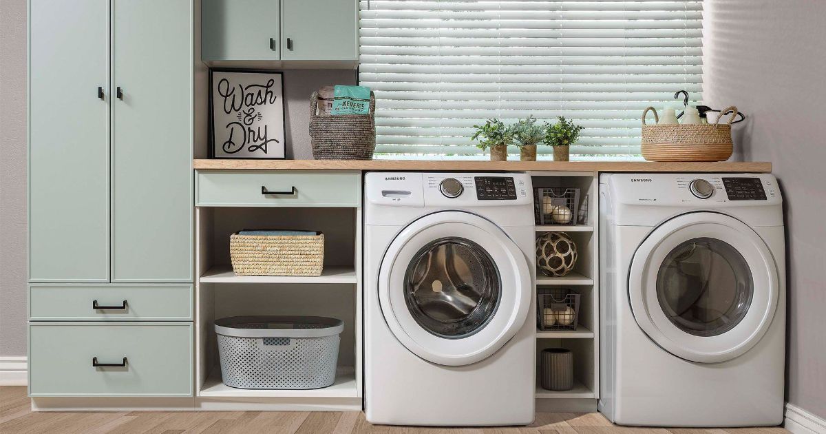 A modern laundry room with light green cabinetry, a washing machine, a dryer, and decorative baskets and plants.