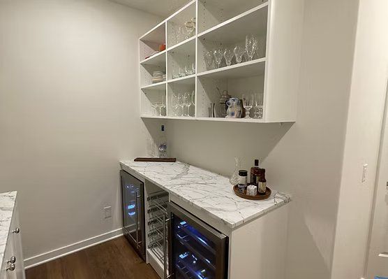 Built-in bar with white cabinets, marble countertop, two wine coolers, and shelves displaying glassware.