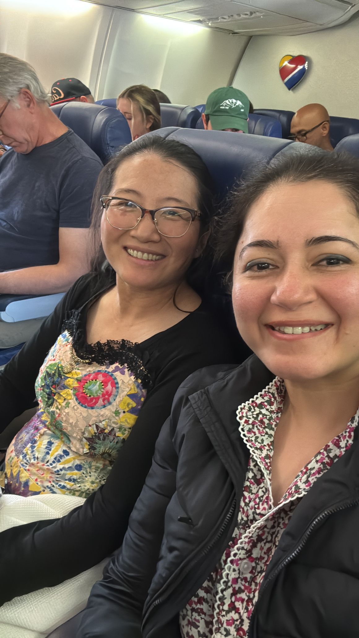 Two smiling passengers seated on an airplane, with cabin seats and windows visible.