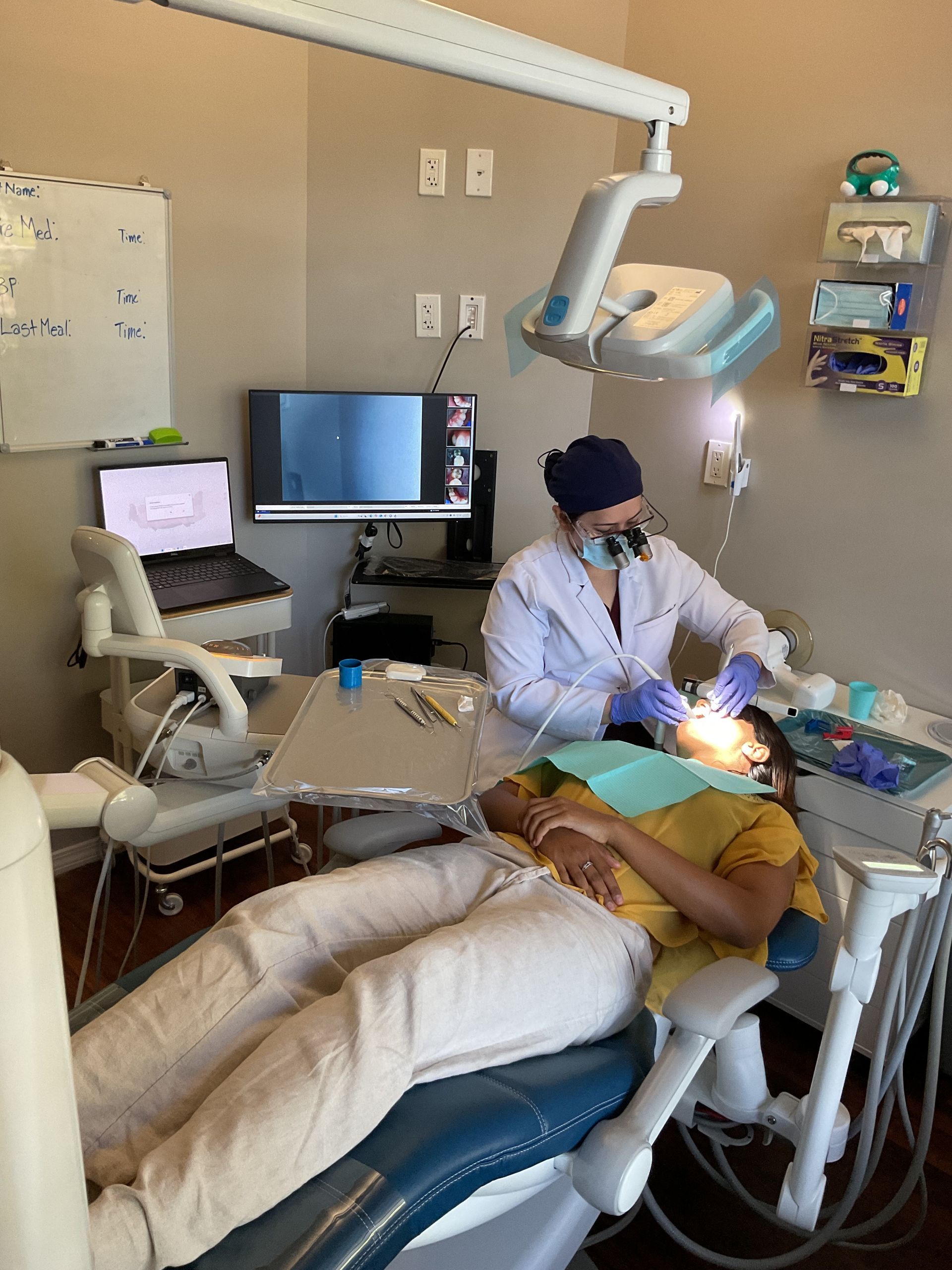 Dentist treating a patient in a dental chair, with bright exam light and monitors in a clinic room.