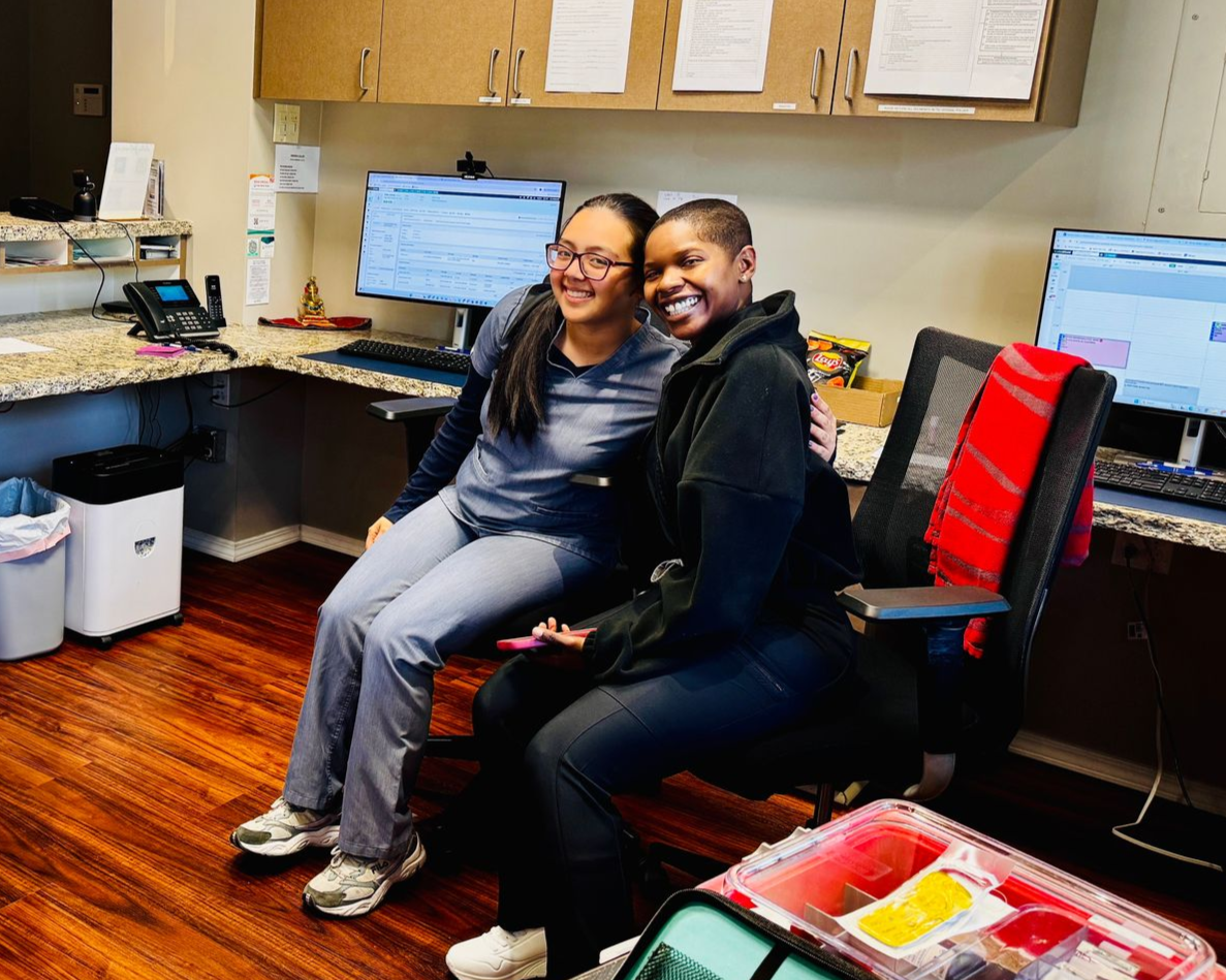Two smiling people seated together in an office with computer monitors and shelves behind them.