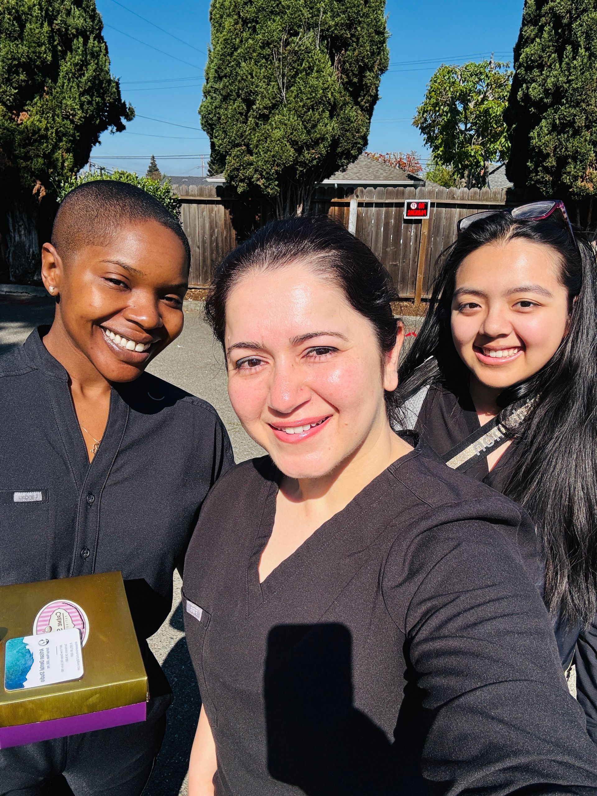 Three people smiling outdoors in bright sunlight, with trees and a clear blue sky behind them.