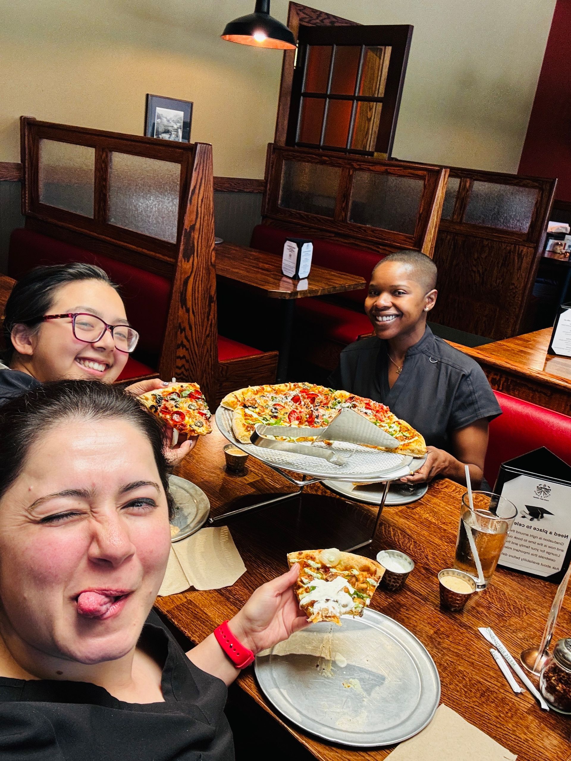Friends smiling at a restaurant table with plates of food and drinks, taking a selfie.