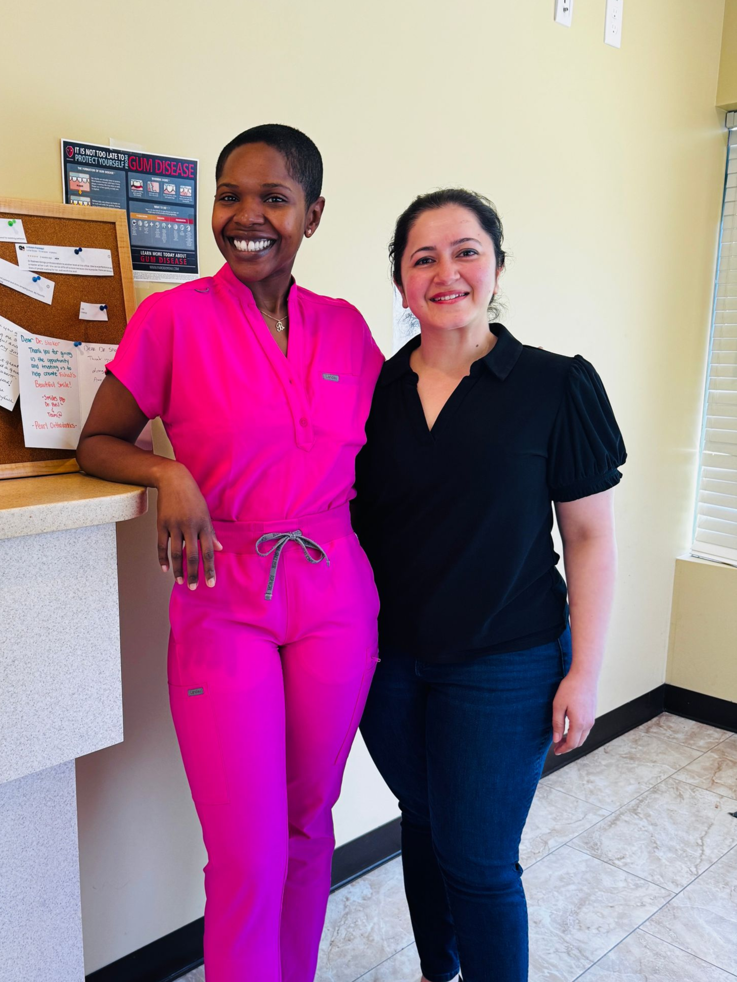 Two people standing together indoors, smiling, one in bright pink and one in black, beside a counter