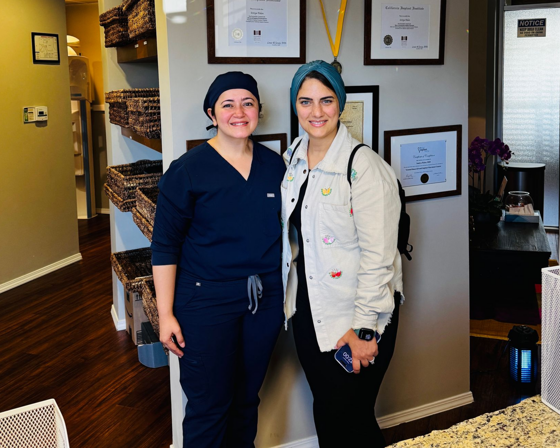 Two smiling coworkers stand in a hallway, one in navy scrubs and one in a white jacket, beside framed certificates.