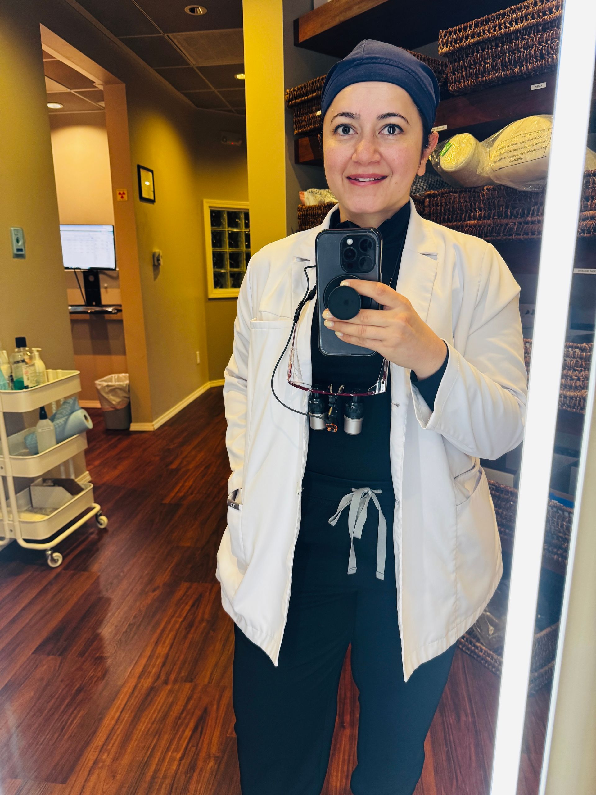 Person taking a mirror selfie in a warmly lit hallway, wearing a white jacket and black outfit.