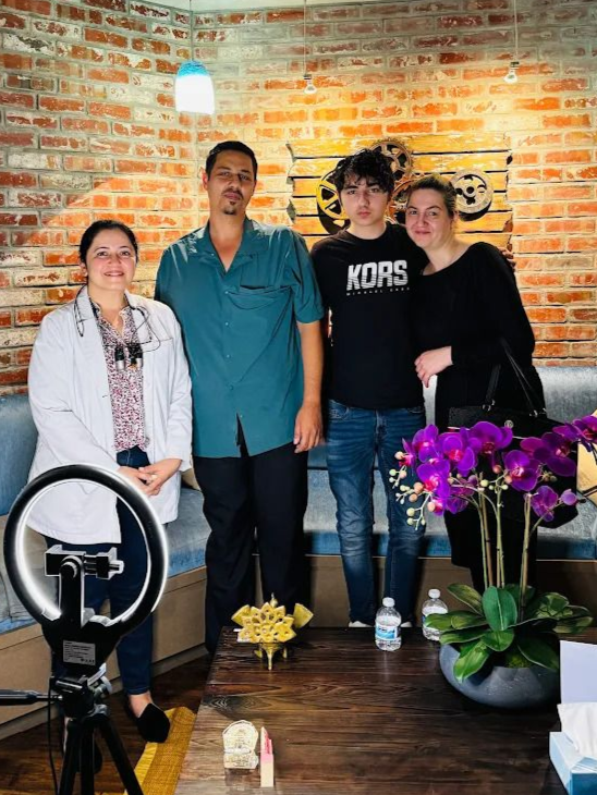 Four people stand together in a room with a brick wall, including a person in a white medical coat with dental loupes.