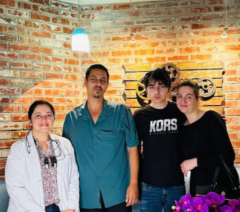 Four people stand together in a room with a brick wall, including a person in a white medical coat with dental loupes.