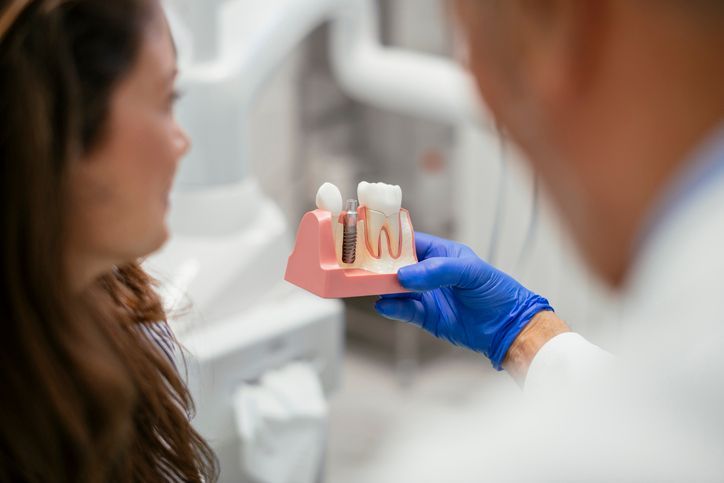 A dentist in blue gloves holds a model showing a dental implant procedure to a patient in a clinical office setting.