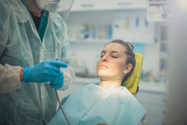 A dental professional in protective gear and blue gloves prepares to treat a patient sitting in a chair.