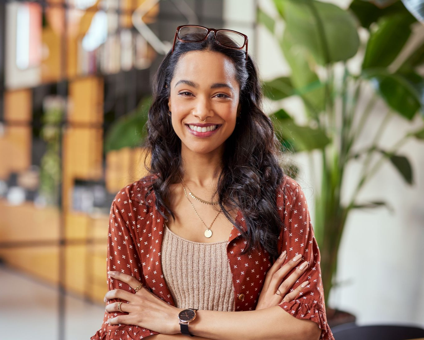 Smiling person with folded arms in a bright indoor setting, wearing a red patterned cardigan and beige top.