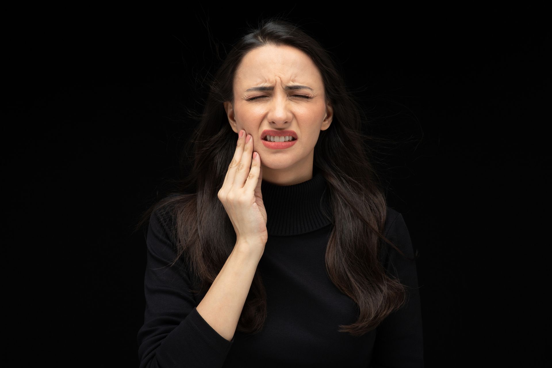 Woman in black turtleneck, holding her cheek in pain, grimacing against a black background.