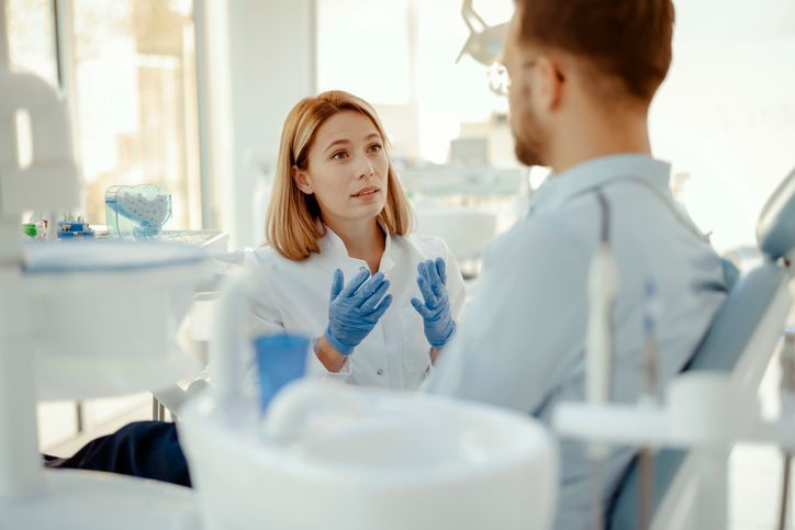 A dentist wearing blue gloves gestures while explaining a procedure to a patient in a dental exam chair.