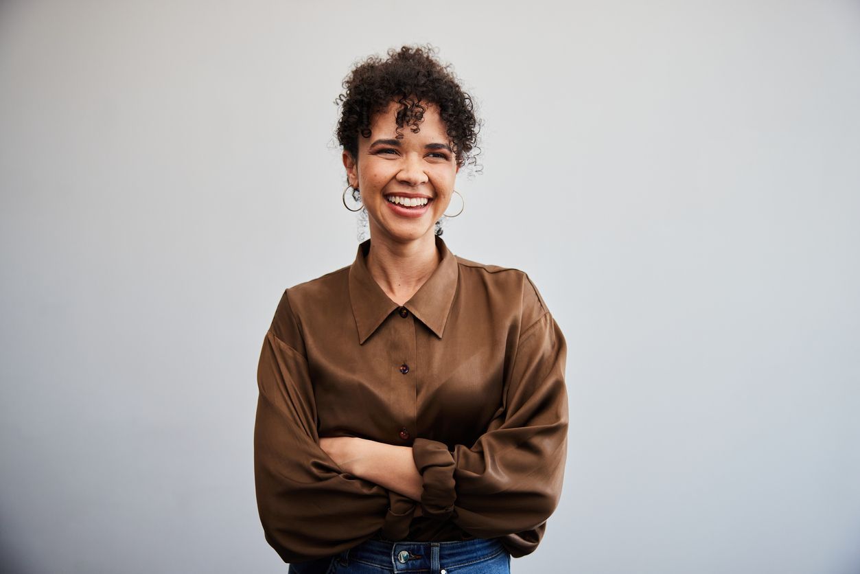 A woman in a brown shirt is standing with her arms crossed and smiling.