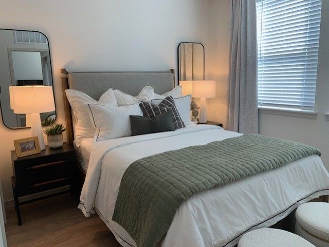 Neatly made bedroom with white bedding, green throw blanket, lamps, and a window with blinds