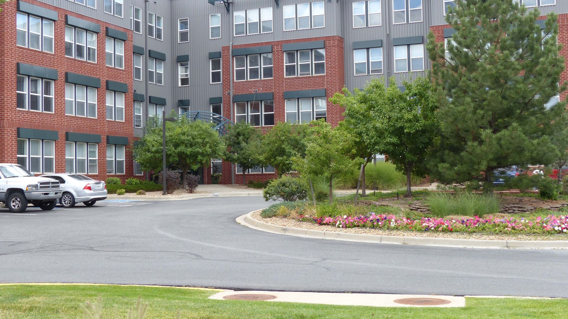 Modern brick apartment building with parked cars, trees, and landscaped circular driveway.