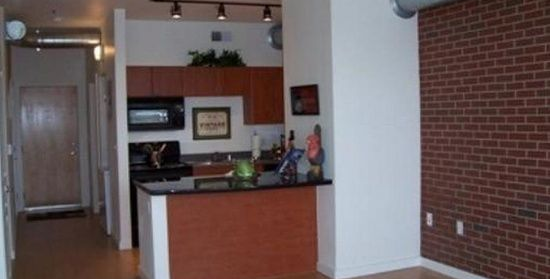 Empty apartment kitchen with red brick accent wall and dark wood cabinets, viewed from the living area