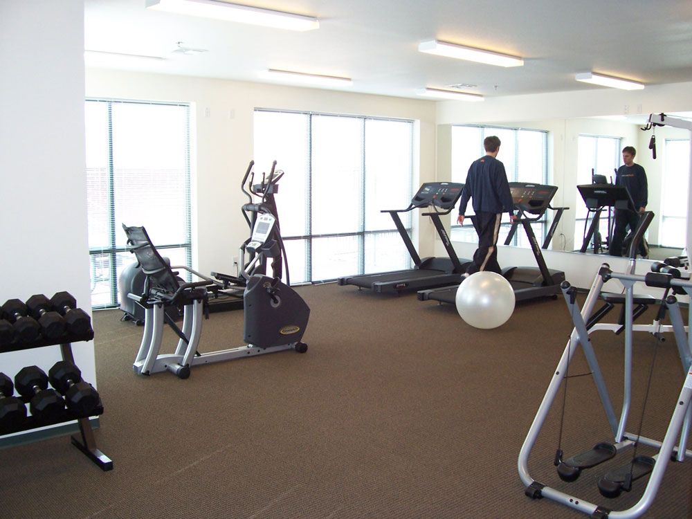 Gym with treadmills, exercise bikes, dumbbells, and two people working out near windows.