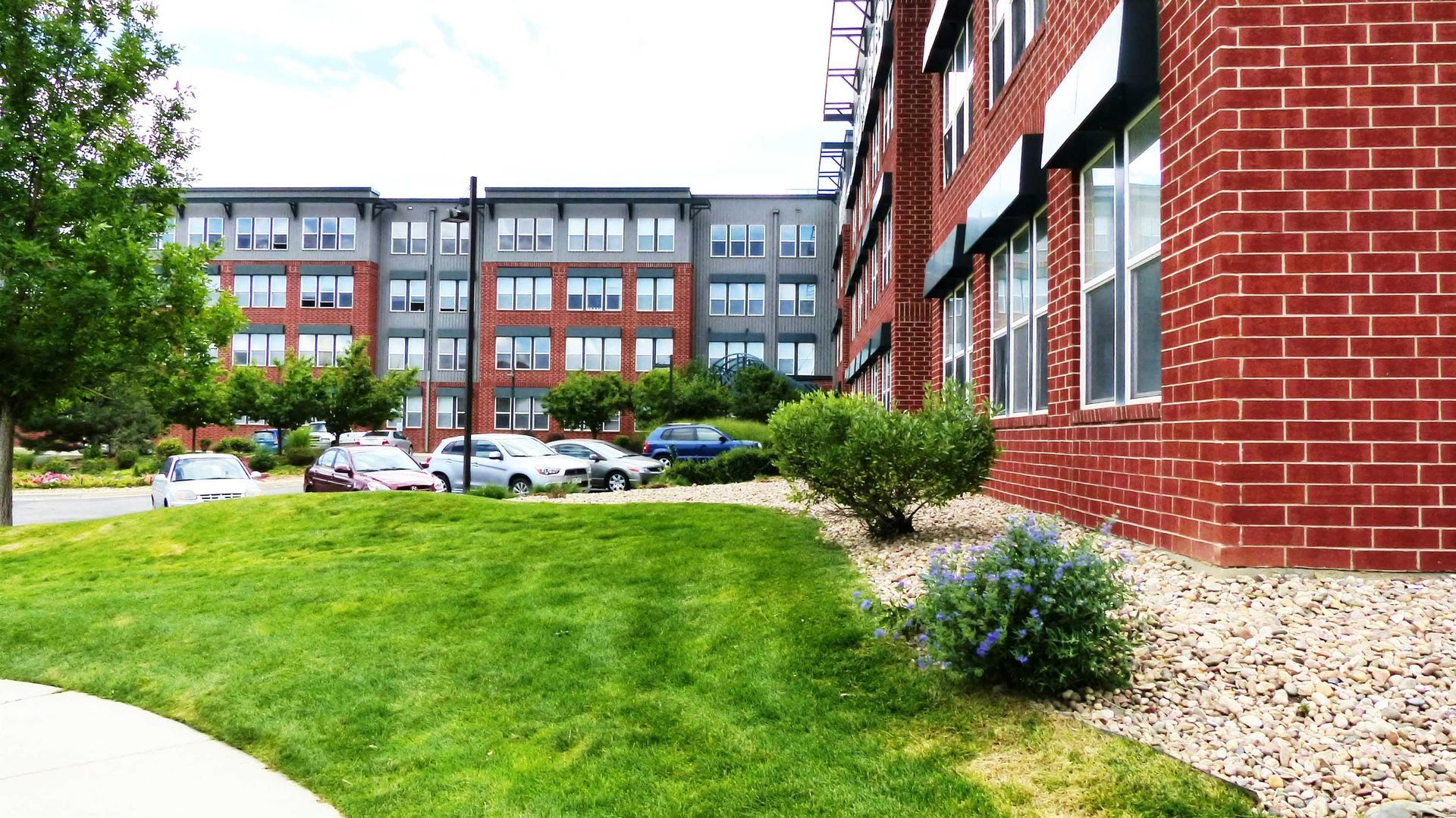 Apartment building courtyard with green lawn, parked cars, and red brick buildings under cloudy sky