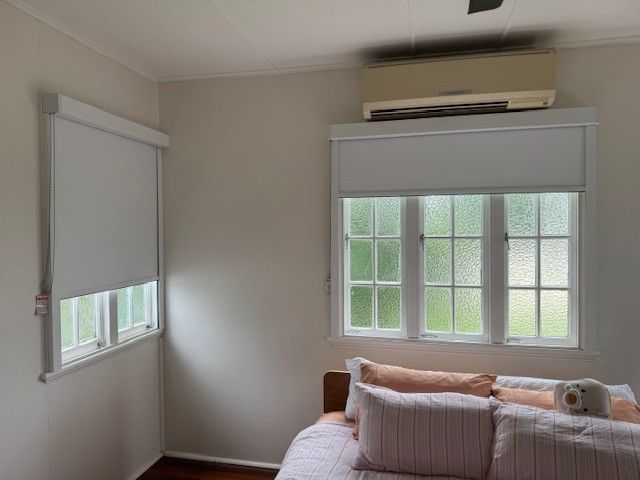 Bright bedroom with two windows, white blinds, and a bed under a wall-mounted air conditioner. — Blinds & Curtains in Townsville, QLD