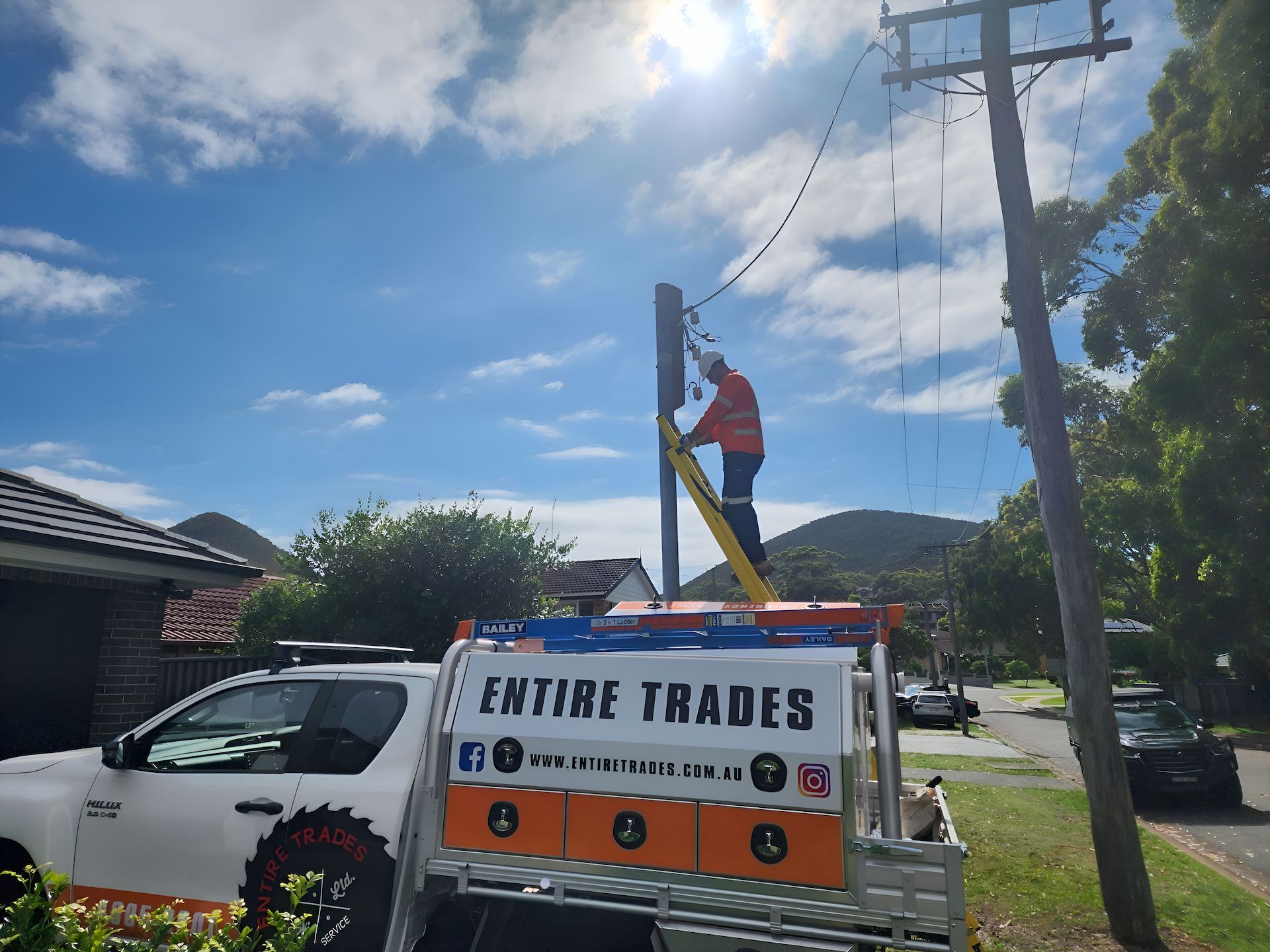 A Man In An Orange Uniform Is Working On A Powerline On A Ladder With A Work Ute In Front — Entire Power Solutions In Maitland, NSW