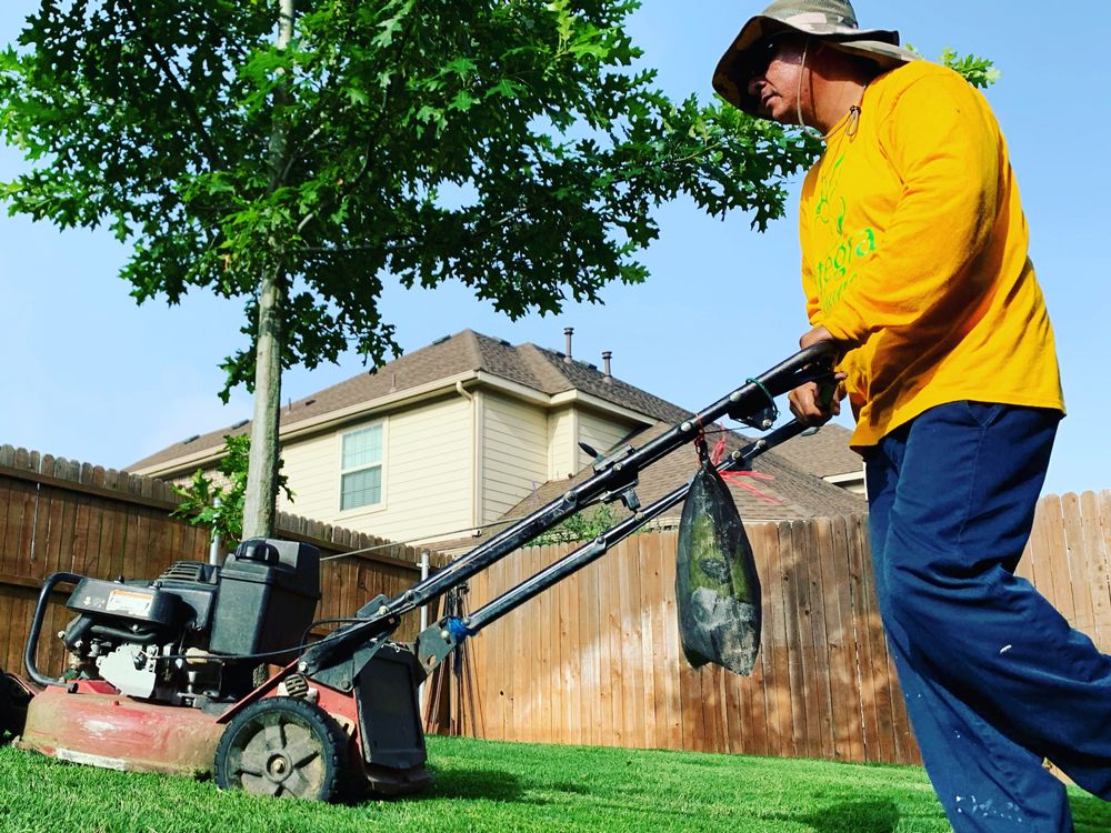 Person mowing lawn in a residential backyard, wearing a yellow shirt, with a house and fence in the background.
