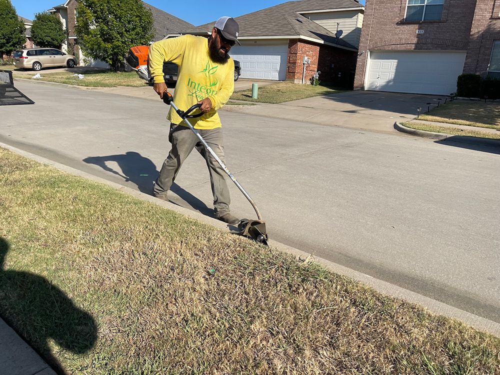 Man with a string trimmer cutting grass along a curb on a sunny day.