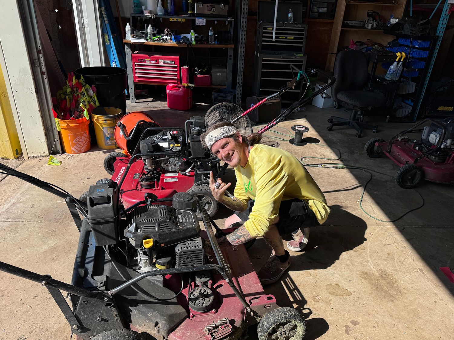 Person crouching near two lawnmowers, smiling, in a garage; tools and shelves in the background.