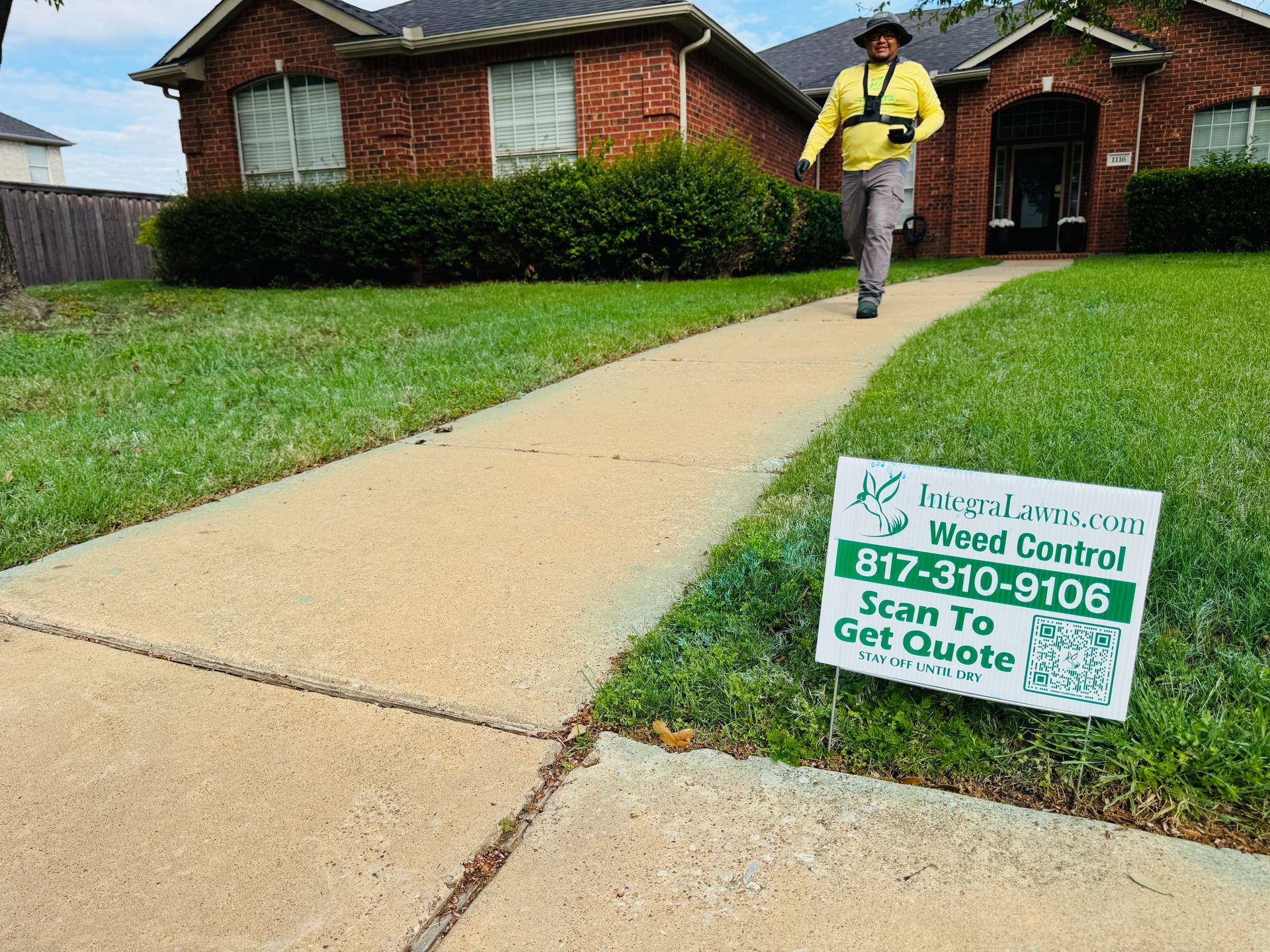 Lawn care worker walking on sidewalk beside brick house; yard sign advertises mowing and lawn services.