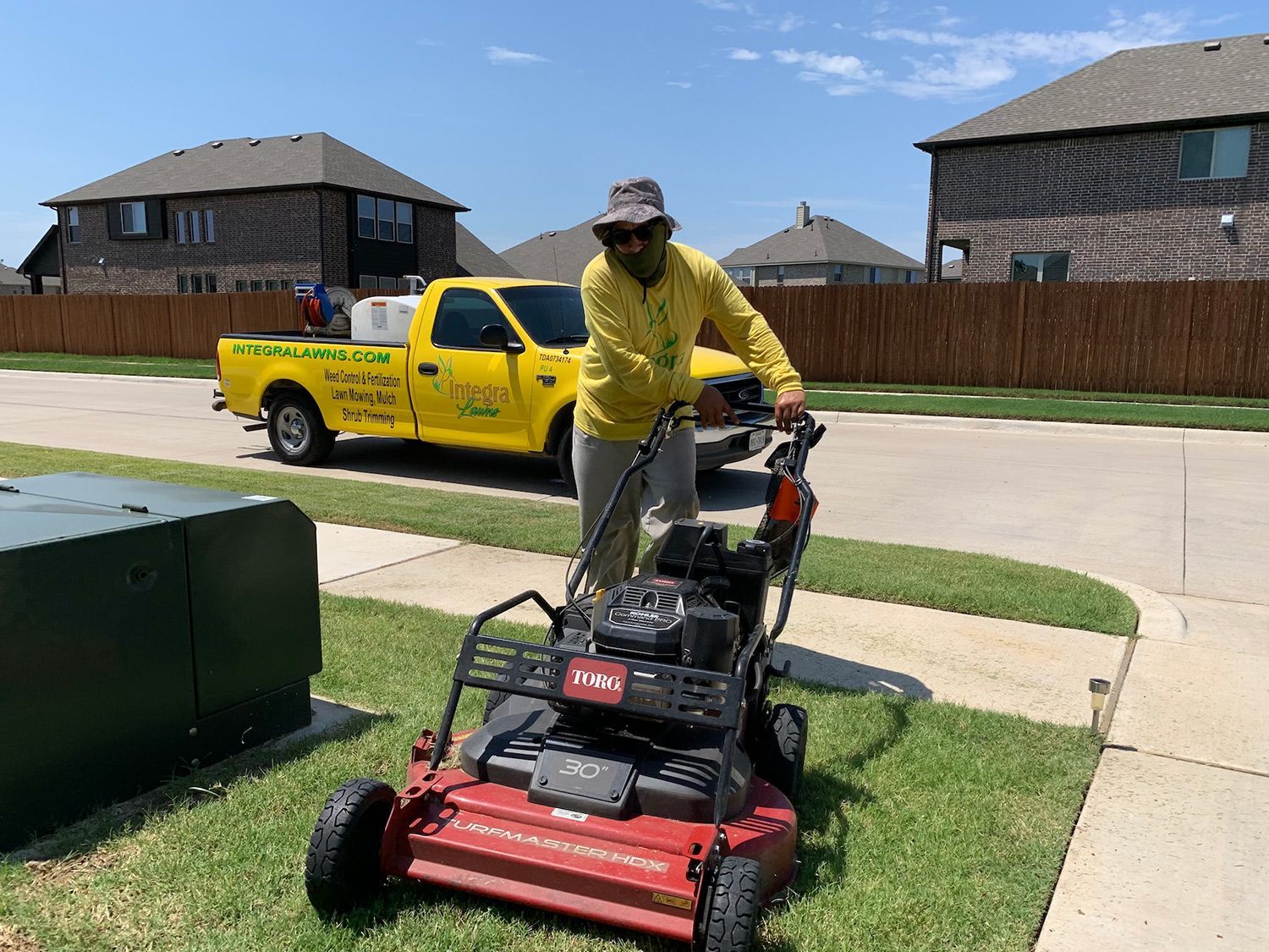Man mowing a lawn with a red and black lawnmower in a residential area. Yellow truck parked nearby.
