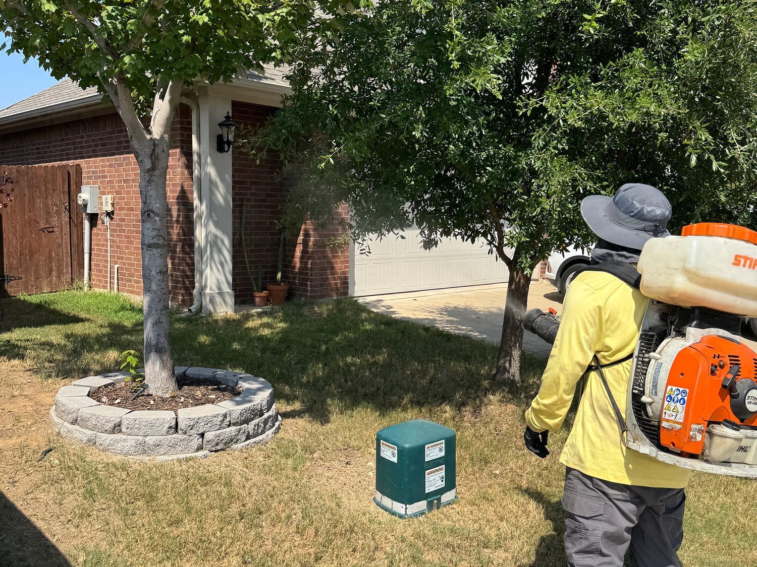 Man spraying a yard with a backpack sprayer near a house and trees; grass and stone border visible.