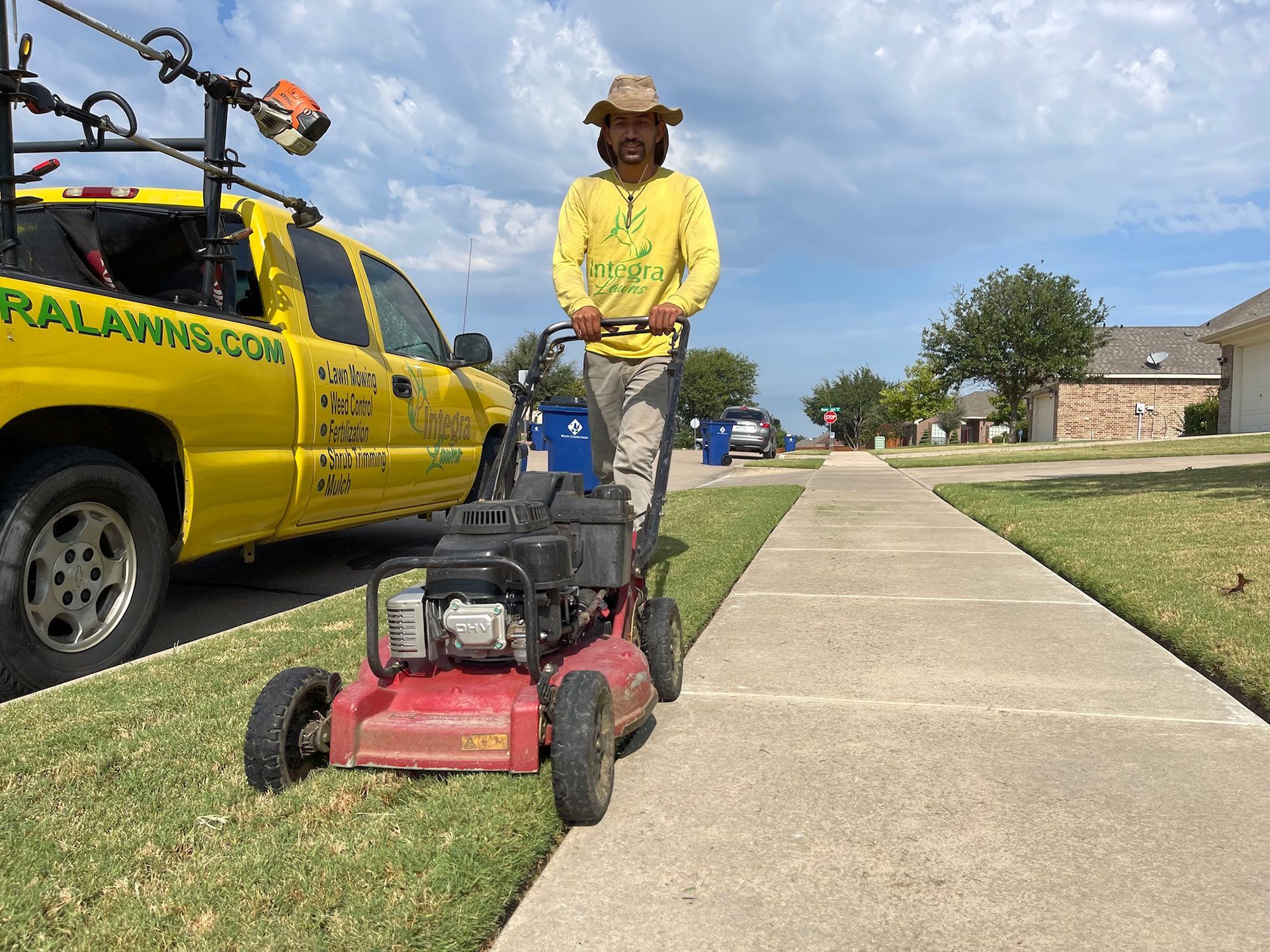 Person mowing grass along a sidewalk with a red lawnmower; yellow truck with logo in background.
