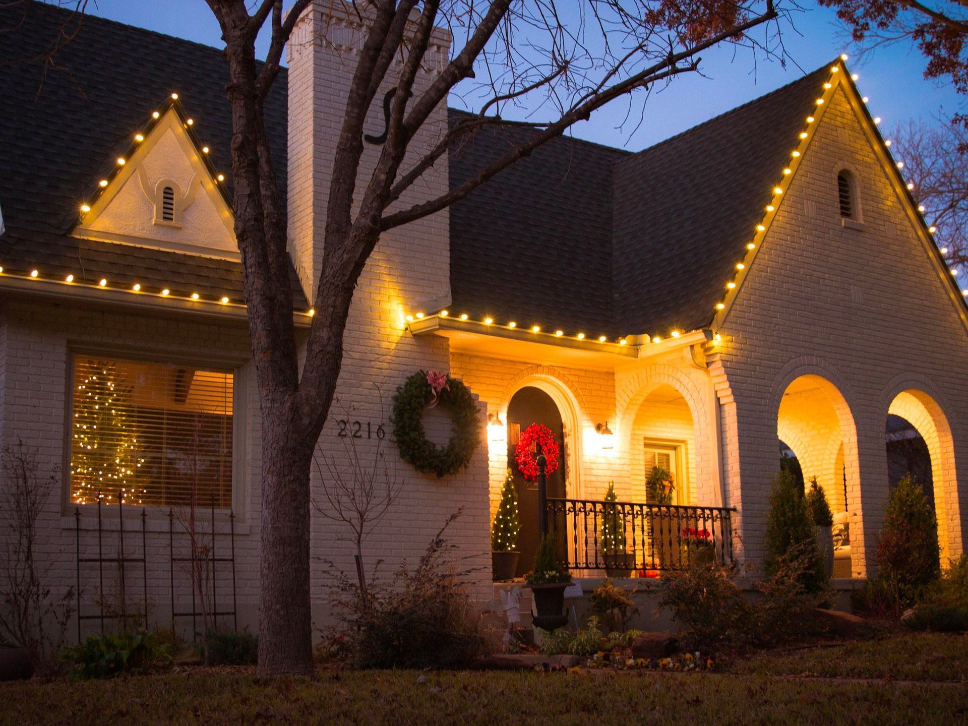 House decorated with warm white Christmas lights, a wreath, and a decorated tree visible in the window.