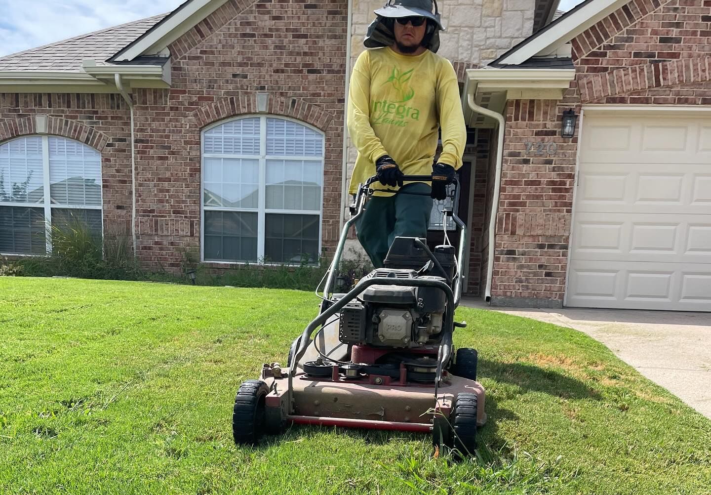 Person mowing lawn with a push mower in front of a brick house with a garage.